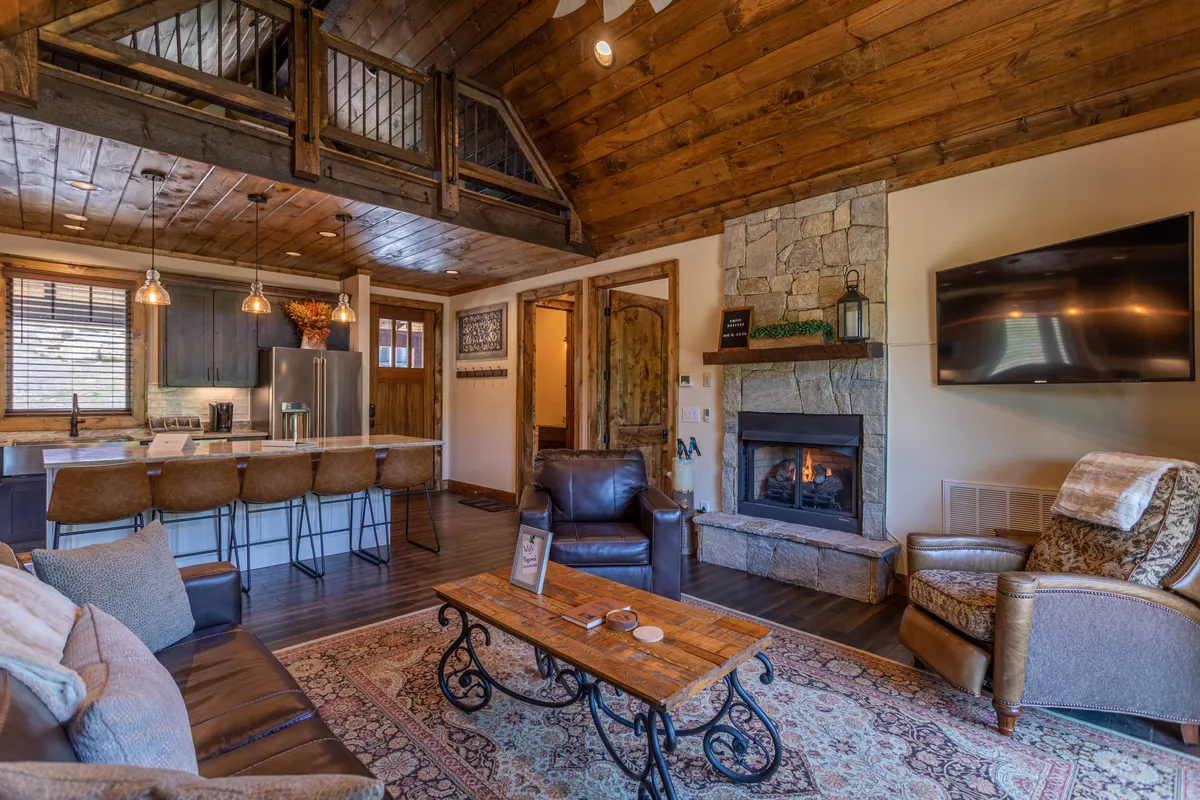 This image showcases the heart of the home, featuring a cozy living area with a fireplace, a plush rug, comfortable sofas, and a kitchen island visible in the background. Exposed wooden beams and the warm glow from the fireplace create a welcoming atmosphere ideal for gatherings or relaxing evenings.