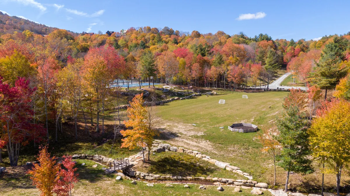 A wide, grassy space framed by vibrant autumn trees and a gentle stream, with a small structure suggesting a play area or picnic spot. The area looks ideal for picnicking, outdoor games, and casual nature exploration.