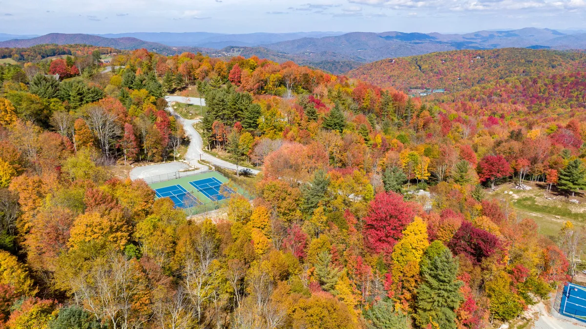 A wide view of tennis courts set amid vibrant autumn foliage with a dramatic mountain range in the background. The courts are surrounded by nature, and the setting sun casts a warm glow across the scene, highlighting the beauty of the outdoor recreational amenities.