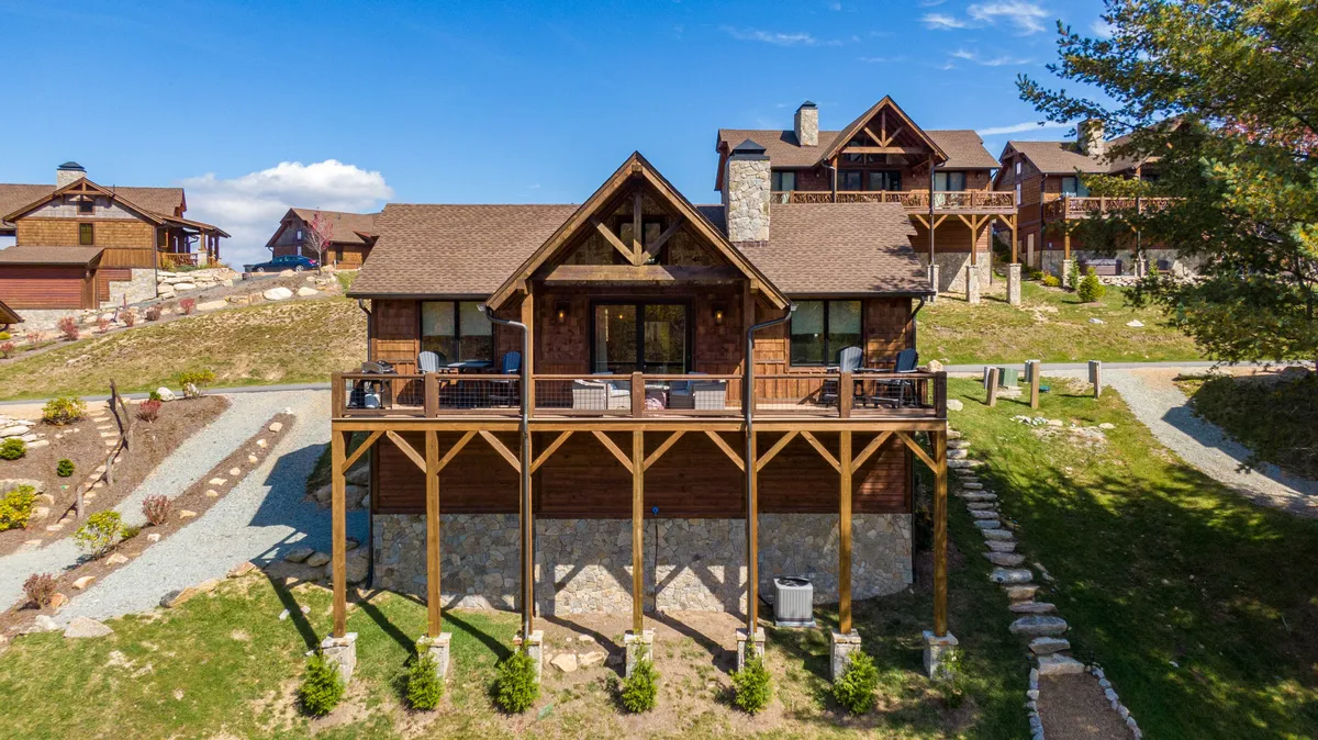 The image shows a charming single-level cabin nestled in lush landscaping, featuring a spacious wooden deck with a stone base. The deck overlooks sweeping mountain views, inviting guests to relax outdoors, enjoy alfresco meals, or simply soak in the serene natural surroundings. The soft evening light adds warmth, highlighting the peaceful mountain retreat vibe.