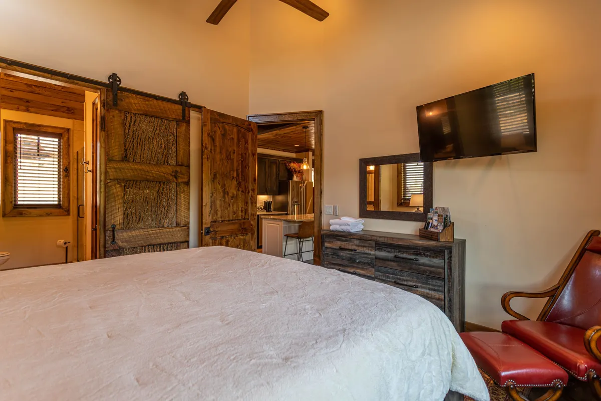 This image shows a comfortable bedroom featuring a bed with white plush bedding, a wooden sliding barn door, a wall-mounted smart TV, a brown wooden dresser, and a red leather chair. Morning sunlight filters in through a window, creating a serene and inviting vibe.