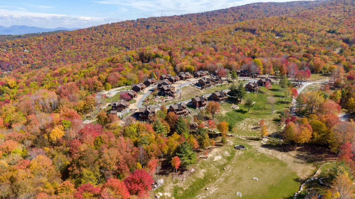 A scenic view featuring colorful autumn foliage, rustic cabins clustered together, and an open green space set against a mountain backdrop. The scene is ideal for hiking, picnicking, and family gatherings, evoking a peaceful and communal atmosphere.