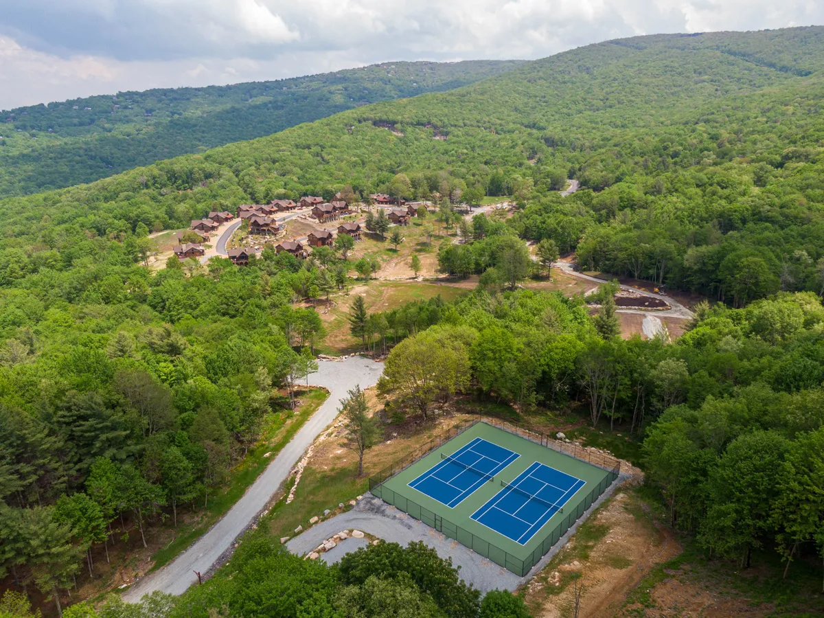 An exterior shot showing tennis courts, a winding road, residential buildings, and the expansive mountain range beyond. The composition captures the relationship between the resort accommodations and the surrounding natural landscape, emphasizing both community and access to the outdoors.