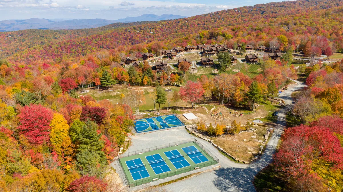 This image captures vibrant fall foliage surrounding tennis courts, with wooden cabins and scenic mountains in the background. Children and families can be imagined enjoying tennis or gathering in this lively, open-air setting.
